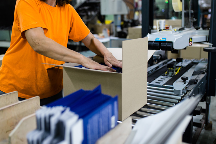 Un ouvrier en t-shirt orange plaçant des produits bleus dans un carton ouvert sur une chaîne de conditionnement, avec d'autres articles en premier plan.