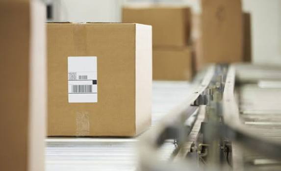 Un carton fermé avec une étiquette de suivi à code-barres posé sur un convoyeur à rouleaux métalliques dans un entrepôt.
