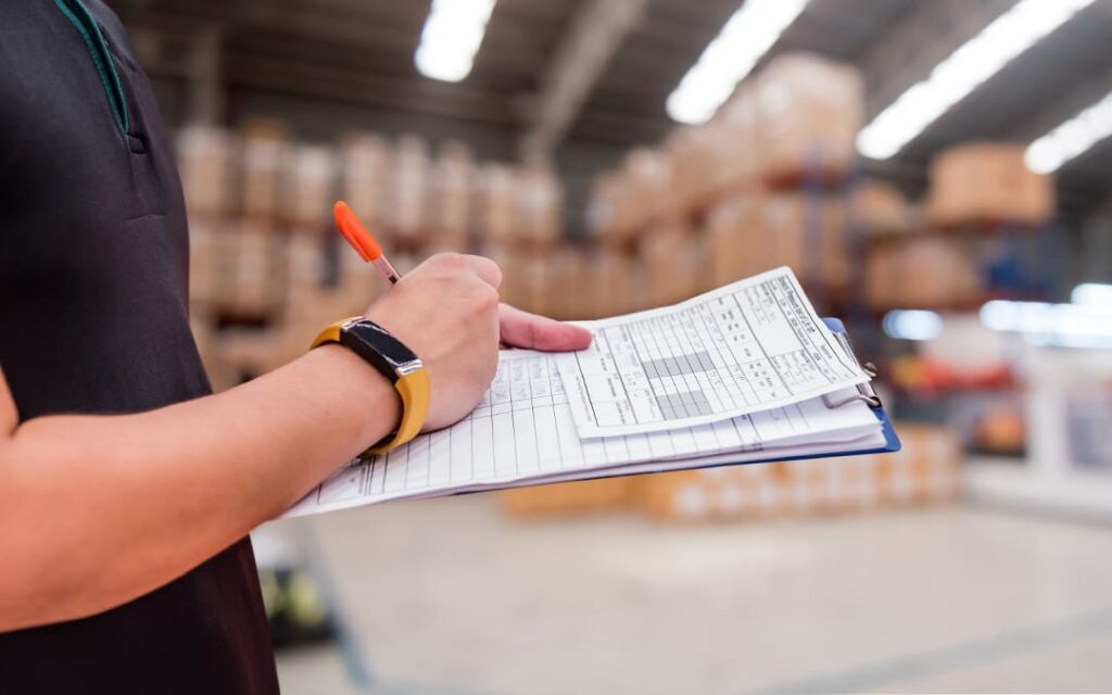 Une personne en uniforme tenant une planche à pince avec un formulaire de contrôle, dans un entrepôt avec des étagères de cartons en arrière-plan.